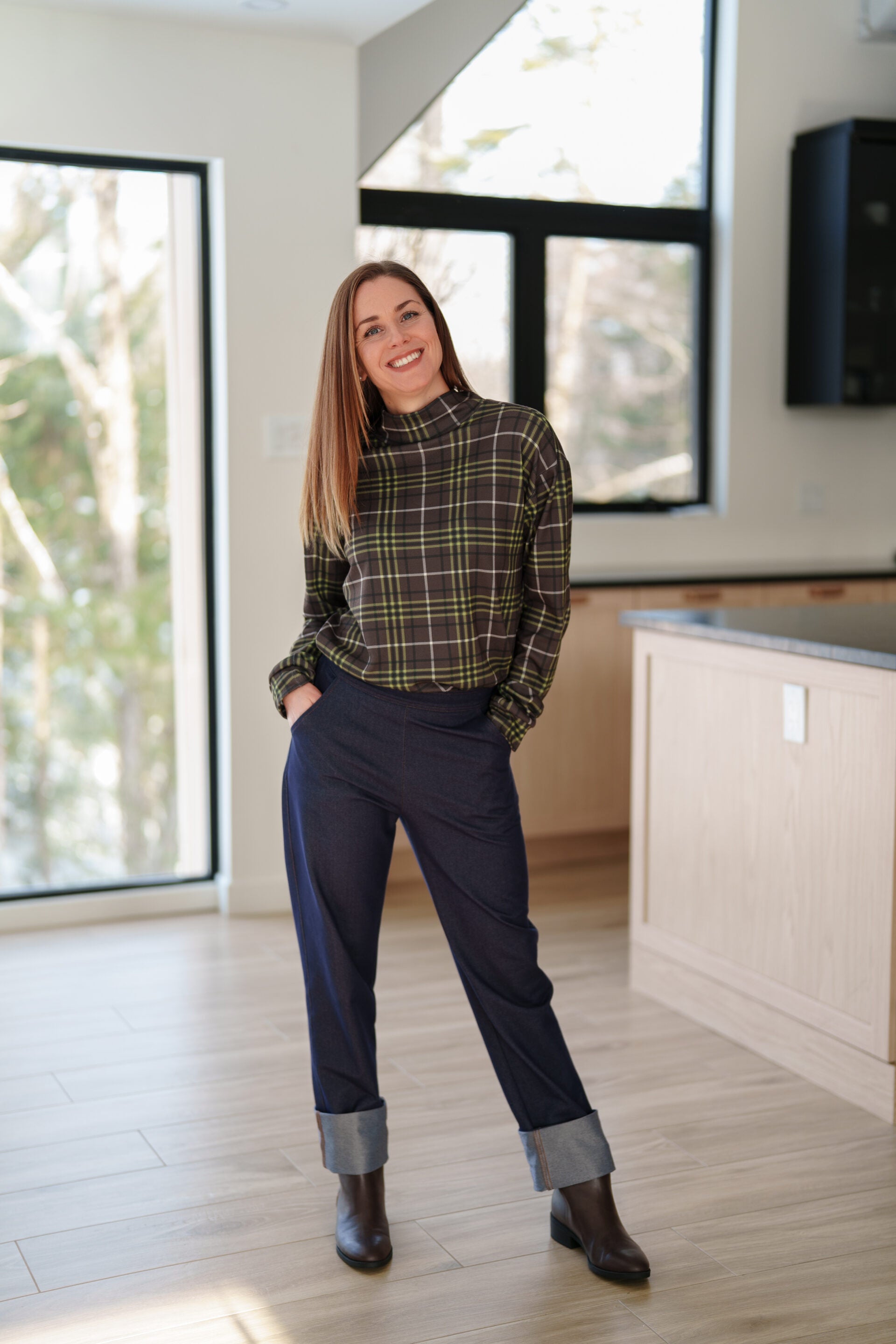 A woman wearing the Yamaska Pants by Rien ne se Perd in Denim Blue, pull on style jeans with rolled cuffs. She is wearing them with a green and brown plaid shirt, and dark brown ankle boots in a bright room with light wood flooring