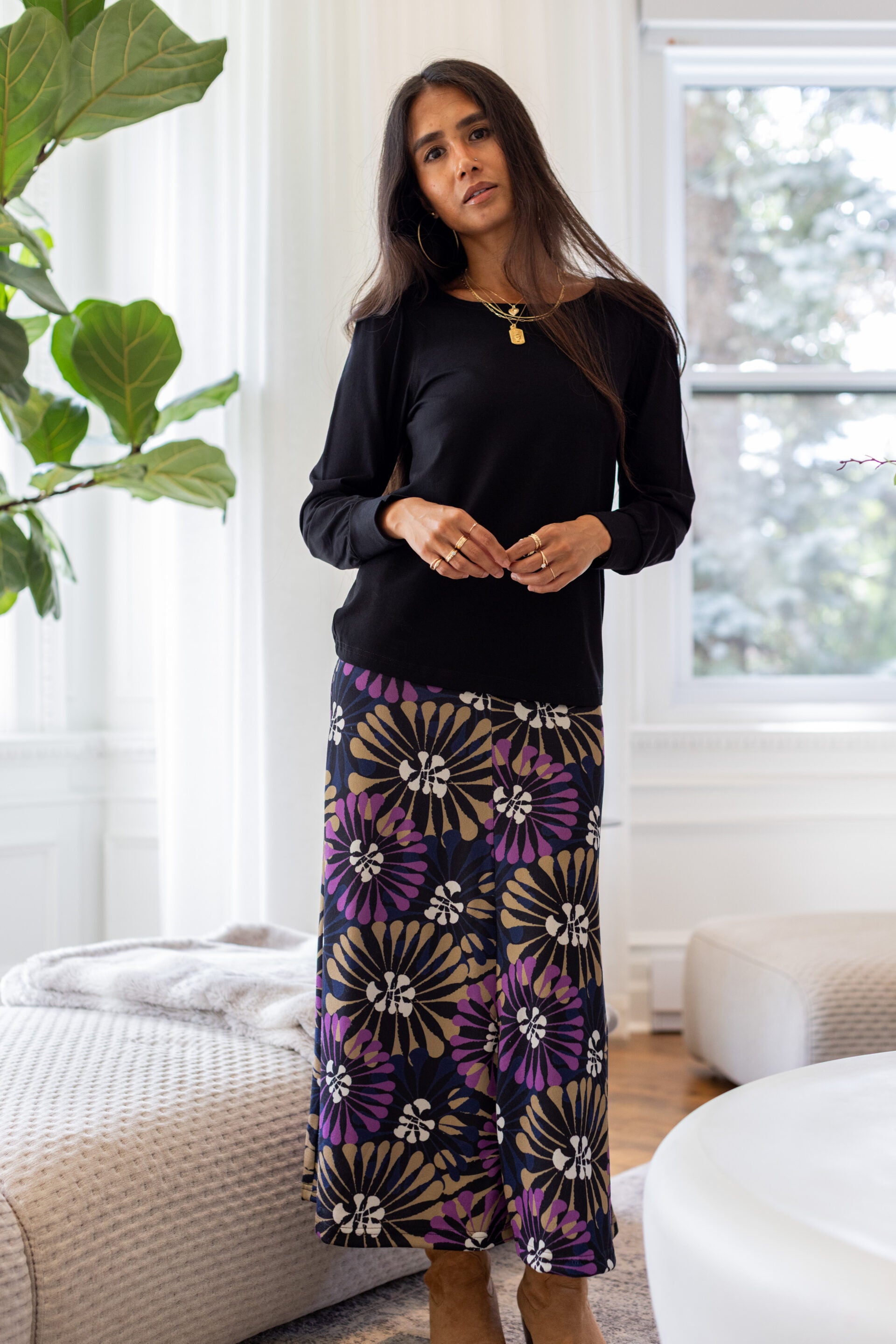A woman standing in a white living room wearing the Becancour Top by Rien ne se Perd in Black, with a round neck and long slightly puffed sleeves with gathered cuffs. She is wearing it with the Hudson Skirt in Floral.
