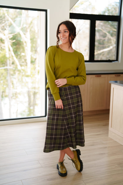 A woman wearing the Becancour Top by Rien ne se Perd in Pistachio, with a round neck and long slightly puffed sleeves with gathered cuffs. She is wearing with a plaid skirt and standing indoors in front of large windows. 
