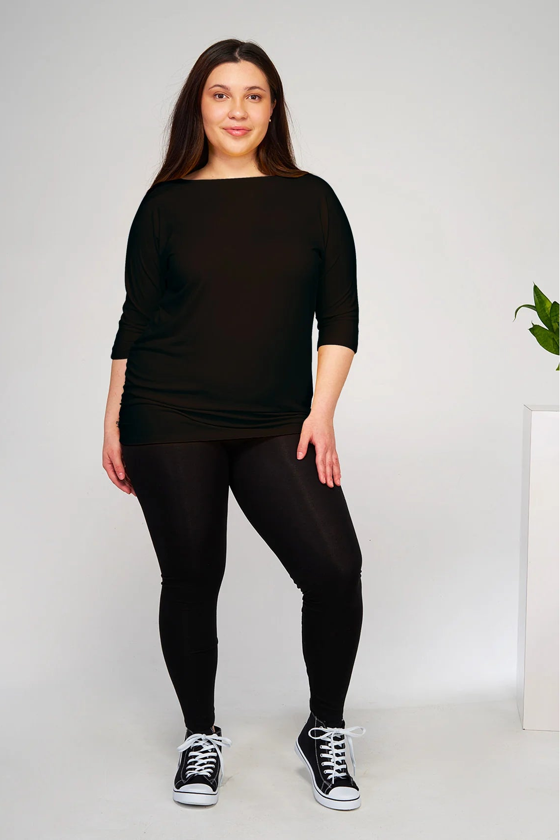 Woman wearing the Bettina top by Moovment in Black with black leggings, standing in front of a white background with a plant. 