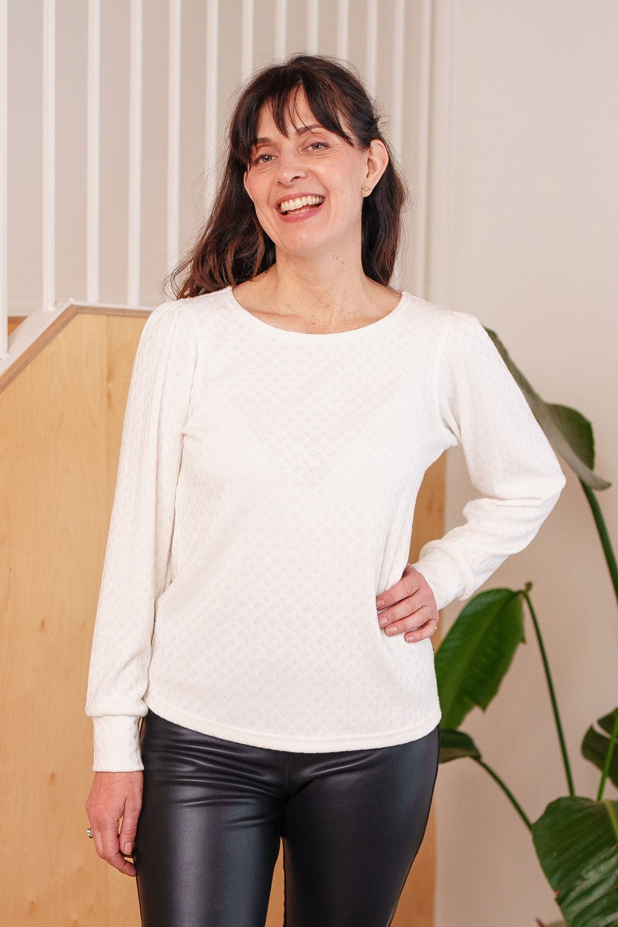 A woman wearing the Camarine Sweater by Rien ne se Perd in Ivory, a cable knit sweater with a scooped neck, puffed shoulders, and gathered cuffs, with a black leather pants. She is standing indoors.
