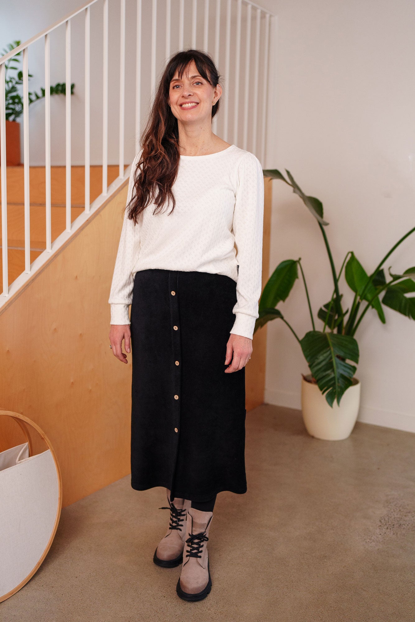 A woman wearing the Camarine Sweater by Rien ne se Perd in Ivory, a cable knit sweater with a scooped neck, puffed shoulders, and gathered cuffs, with a black skirt. She is standing indoors.