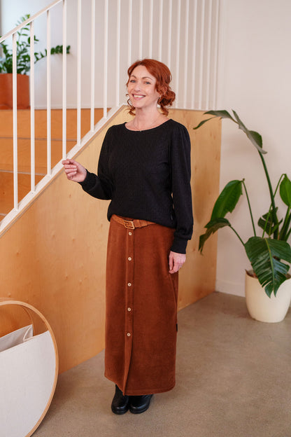 A woman wearing the Camarine Sweater by Rien ne se Perd in Black, a cable knit sweater with a scooped neck, puffed shoulders, and gathered cuffs, with a copper skirt She is standing indoors by a staircase.