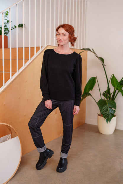 A woman wearing the Cypres Pants by Rien ne se Perd in Blue Denim, stretchy faux denim pants with a pull on waist and tone-on-tone patches on the knees. She is wearing with them with a black sweater and standing in front of a railing. 