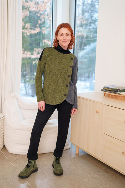 A woman wearing the Lichen Pant by Rien ne se Perd in Black, skinny leg pull-on pants in stretchy corduroy fabric. She is wearing them with green and grey top and standing in front of a window. 