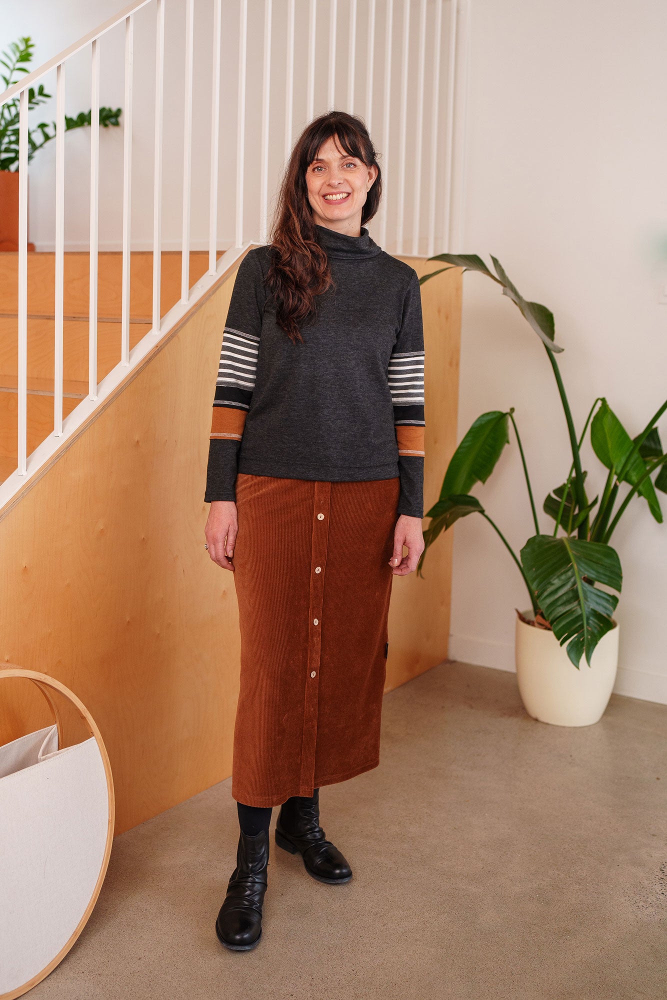 A woman wearing the Chicouté Sweater by Rien ne se Perd in Charcoal/Cognac, with a small turtleneck colour-blocked sleeves. She is wearing it with the Pruche skirt in Cognac and standing in front of a railing. 