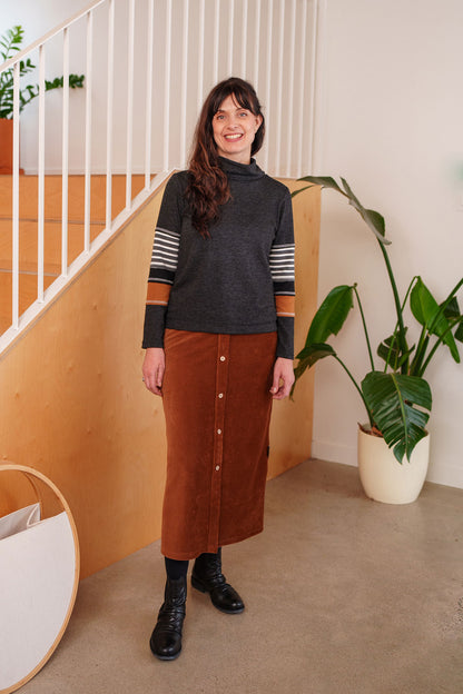 A woman wearing the Chicouté Sweater by Rien ne se Perd in Charcoal/Cognac, with a small turtleneck colour-blocked sleeves. She is wearing it with the Pruche skirt in Cognac and standing in front of a railing. 