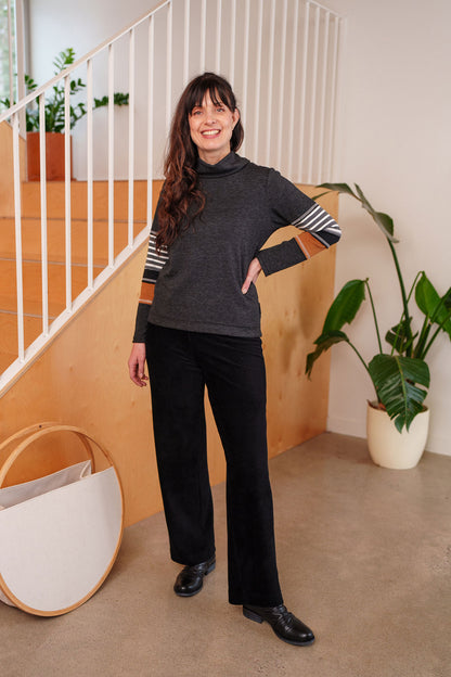 A woman wearing the Chicouté Sweater by Rien ne se Perd in Charcoal/Cognac, with a small turtleneck colour-blocked sleeves. She is wearing it with black pants and standing in front of a railing. 