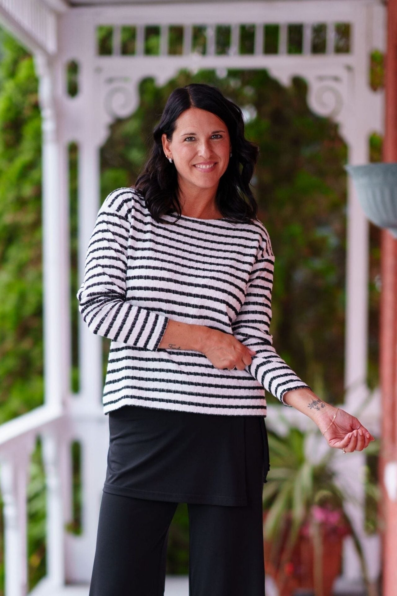 A woman wearing the Collins Reversible Sweater by Rien ne se Perd in Black/White. It's reversible back to front with stripes on side and solid fabric on the other. It has long striped sleeves and a round neck. She is wearing it with black bottoms and standing on a porch with plants. 