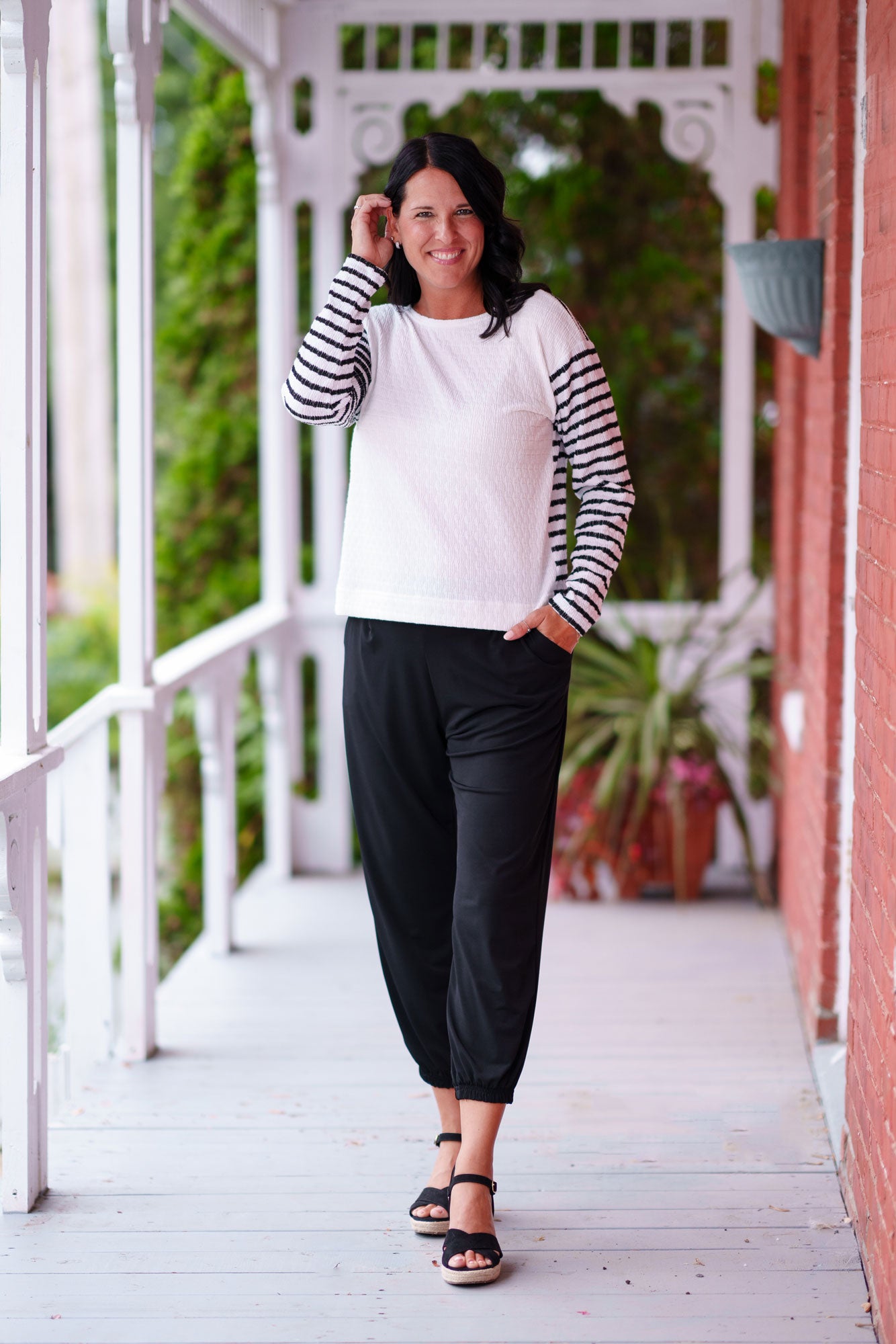A woman wearing the Collins Reversible Sweater by Rien ne se Perd in Black/White. It's reversible back to front with stripes on side and solid fabric on the other. It has long striped sleeves and a round neck. She is wearing with black pants and standing on a porch. 