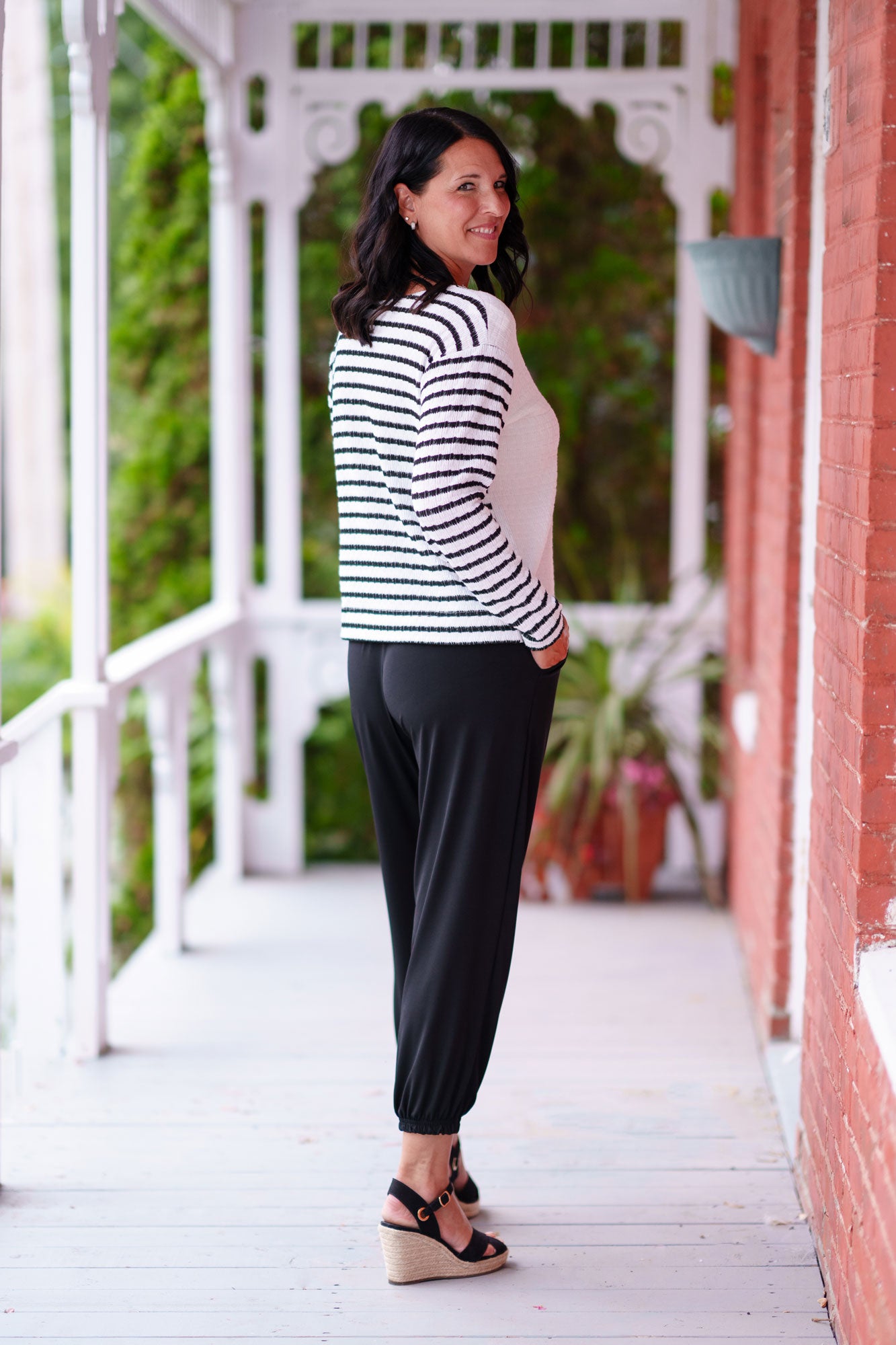 Back view of a woman wearing the Collins Reversible Sweater by Rien ne se Perd in Black/White. It's reversible back to front with stripes on side and solid fabric on the other. It has long striped sleeves and a round neck. She is wearing with black pants and standing on a porch. 