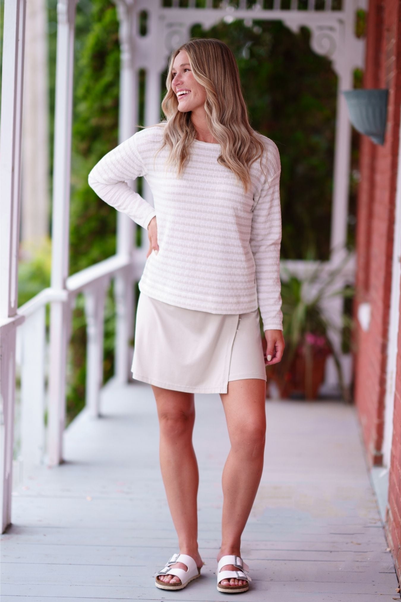 A woman wearing the Collins Reversible Sweater by Rien ne se Perd in Sand/White. It's reversible back to front with stripes on side and solid fabric on the other. It has long striped sleeves and a round neck. She is wearing it with a beige skirt and standing on a porch. 