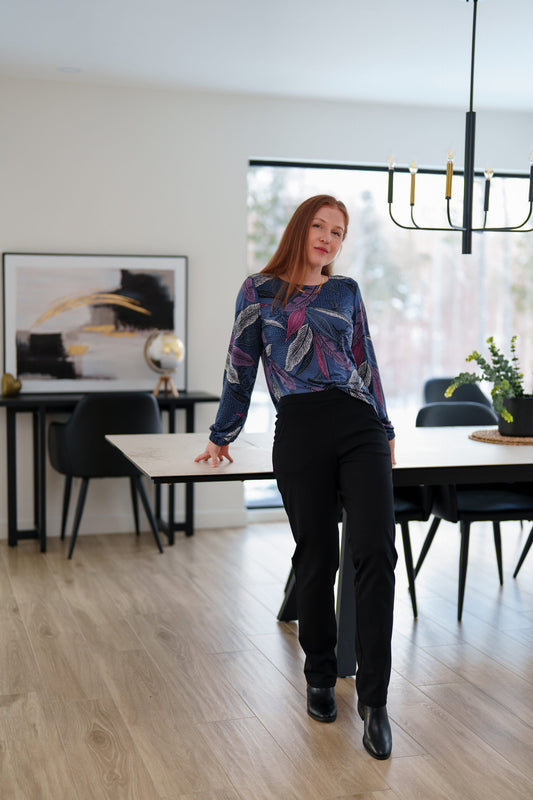 A woman wearing the St Charles Sweater by Rien ne se Perd in Foliage, a jewel toned patterned top with long sleeves and elastic cuffs. She is wearing it with the Murphy Pants in Black and 
standing in a modern kitchen. 
