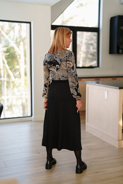 Back view of a woman wearing the Memphre Skirt from Rien ne se perd in Black, a long skirt made from a stretchy pleated fabric that gives a corduroy look. She is wearing it with a paisley top and standing in a house. 