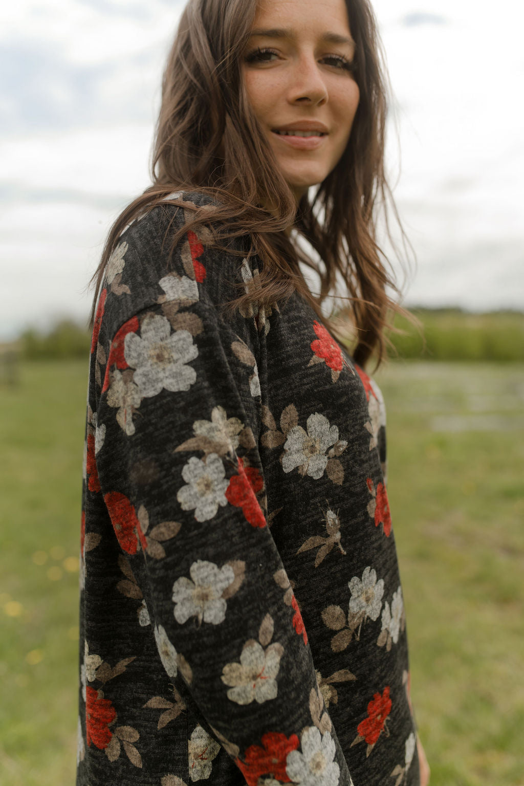 Side view of a woman wearing the Leran Sweater by Rien ne se Perd in Floral print, a mock-neck knit sweater with a black, white and red pattern. She is wearing it with jeans and standing outside in a field. 