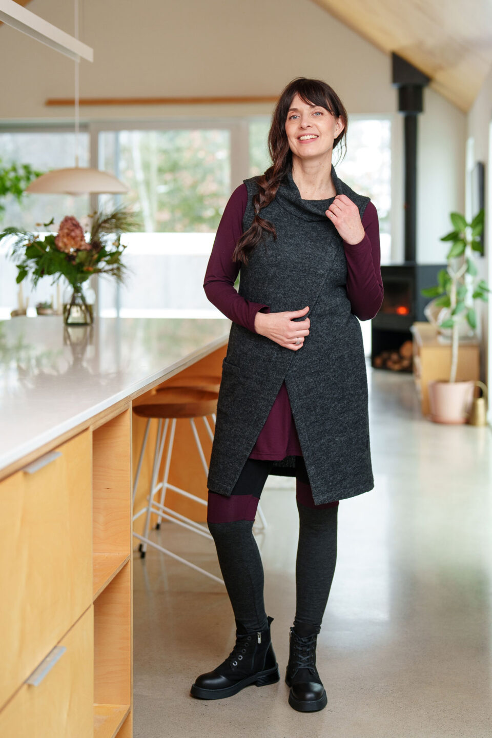 A woman wearing the long Baumier Vest by Rien ne se Perd in Charcoal, with a cowl neck, overlapping front, buttons and the shoulder and a patch pocket. She is wearing it over a burgundy top and leggings and standing in a kitchen. 