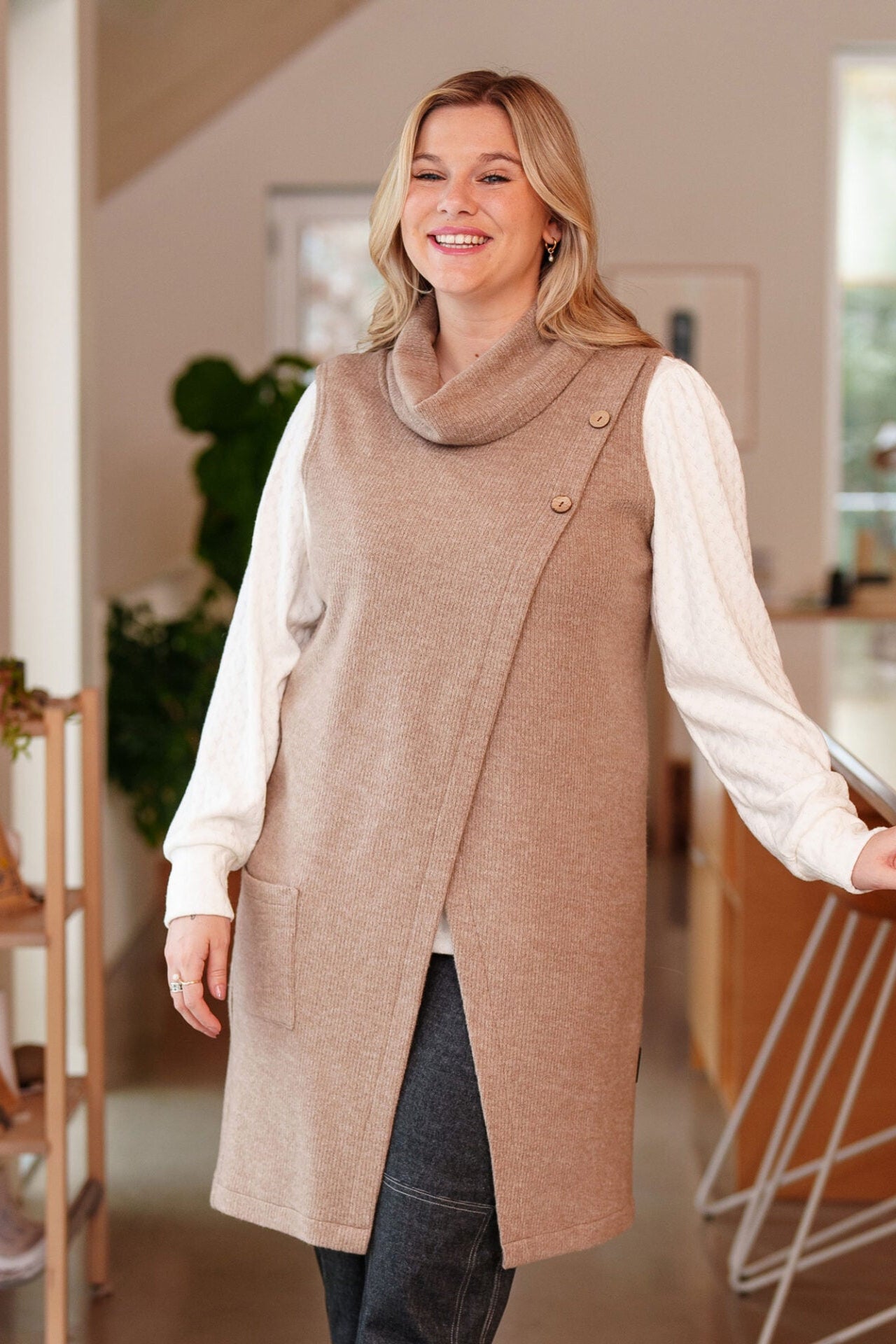 A woman wearing the long Baumier Vest by Rien ne se Perd in Natural, with a cowl neck, overlapping front, buttons and the shoulder and a patch pocket. She is wearing it over a white top and with denim pants and standing in a kitchen. 