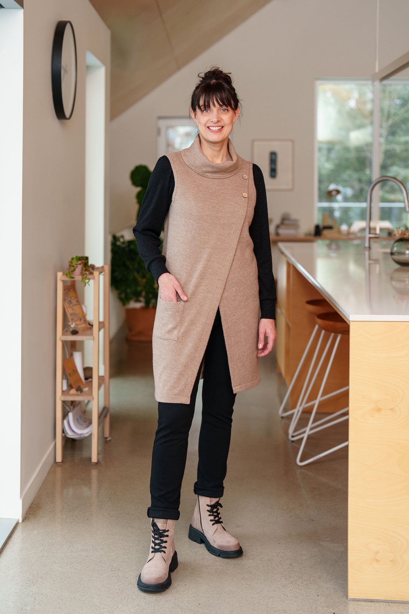 A woman wearing the long Baumier Vest by Rien ne se Perd in Natural, with a cowl neck, overlapping front, buttons and the shoulder and a patch pocket. She is wearing it over a black top and pants and standing in a kitchen. 