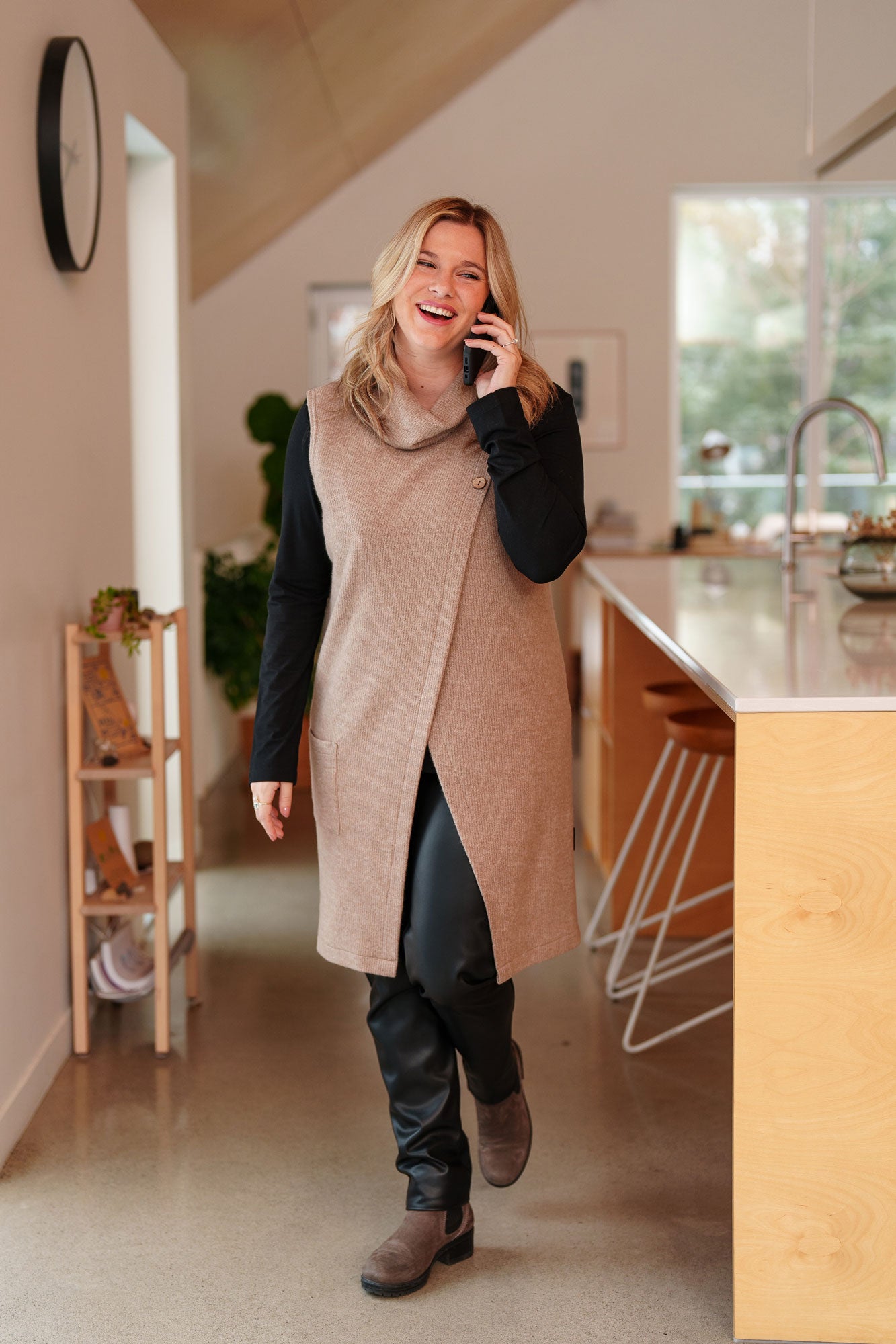 A woman wearing the long Baumier Vest by Rien ne se Perd in Natural, with a cowl neck, overlapping front, buttons and the shoulder and a patch pocket. She is wearing it over a black top and the Resinosa Pants and standing in a kitchen.
