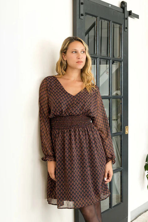 A woman wearing the Spritz Dress by Cherry Bobin in Brown Pattern, featuring puff sleeves, a v-neck,  cinch waisted, and a full knee-length skirt. She is standing in a white room with a black staircase. 