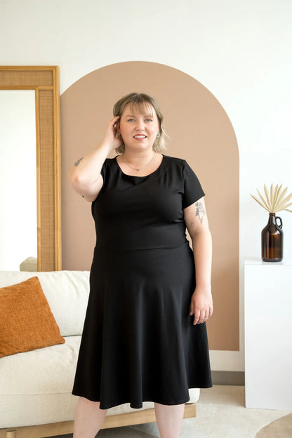 A woman wearing the Bellini Dress by Cherry Bobin in Black, with short sleeves, a round neck, and a fit and flare shape that falls to the knee. She is standing in a room with a couch in it.