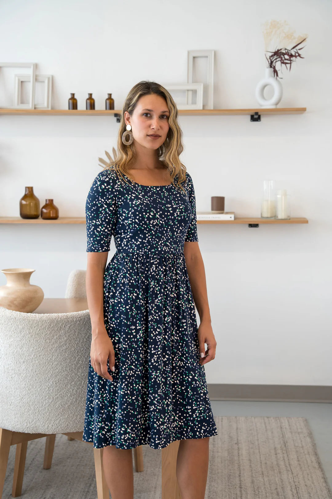 A woman wearing the Doppio Dress by Cherry Bobin in Multicolour Dots, with slight scooped neck, half sleeves, a fit and flare shape, and full skirt that falls below the knee. She is standing in a white room with wooden shelves. 