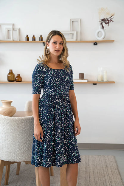 A woman wearing the Doppio Dress by Cherry Bobin in Multicolour Dots, with slight scooped neck, half sleeves, a fit and flare shape, and full skirt that falls below the knee. She is standing in a white room with wooden shelves. 