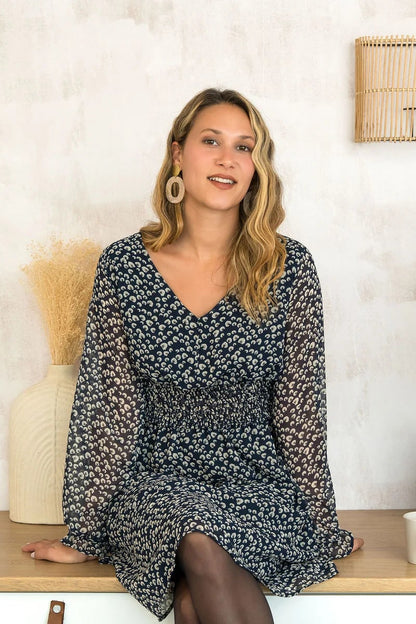 A woman wearing the Spritz Dress by Cherry Bobin in Blue Pattern, featuring puff sleeves, a v-neck,  cinch waisted, and a full knee-length skirt. She is sitting on a wooden counter. 