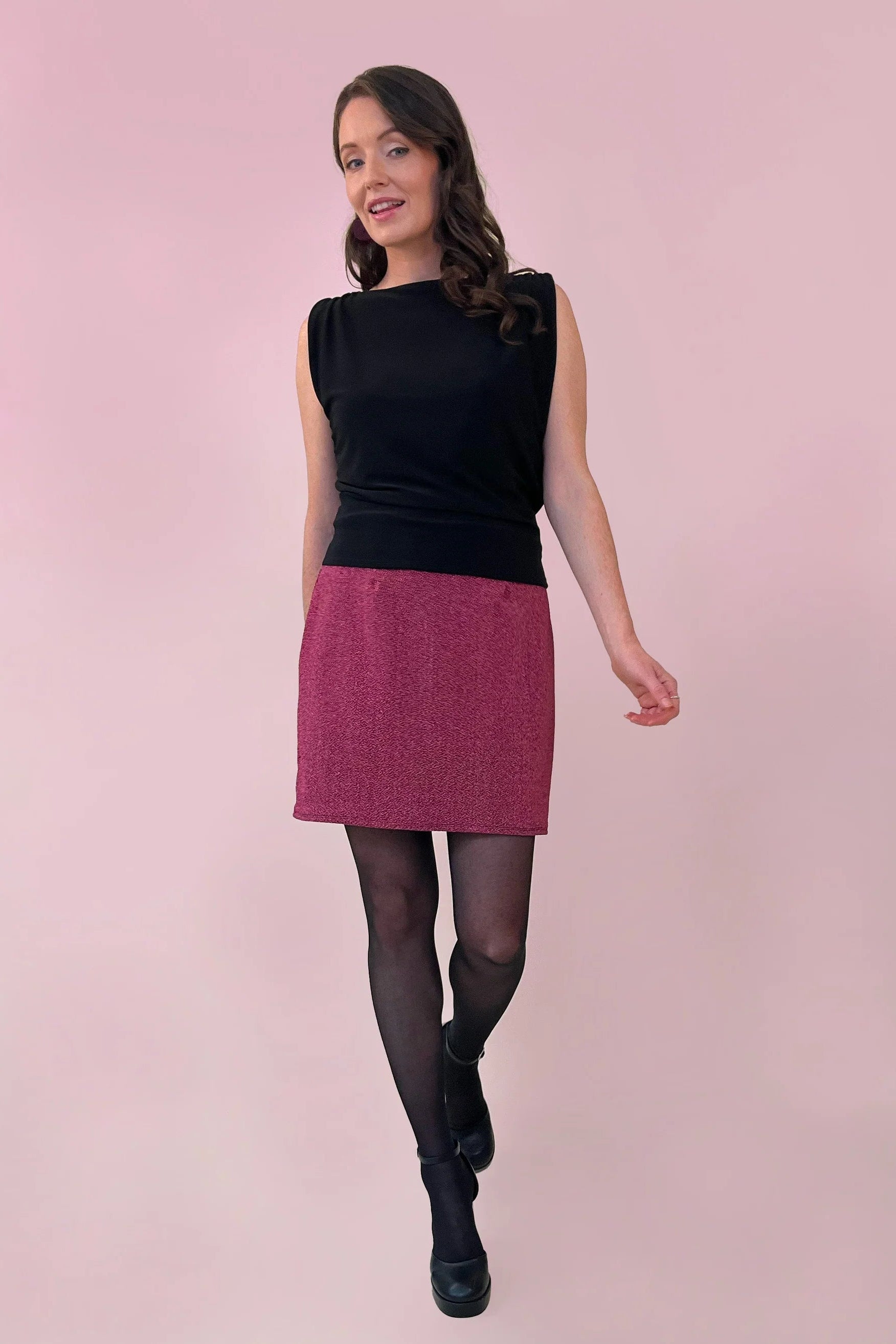 A woman wearing the Glitter Skirt by Canopée in Pink, a short stretchy skirt She's wearing it with a black sleeveless top against a pink background.