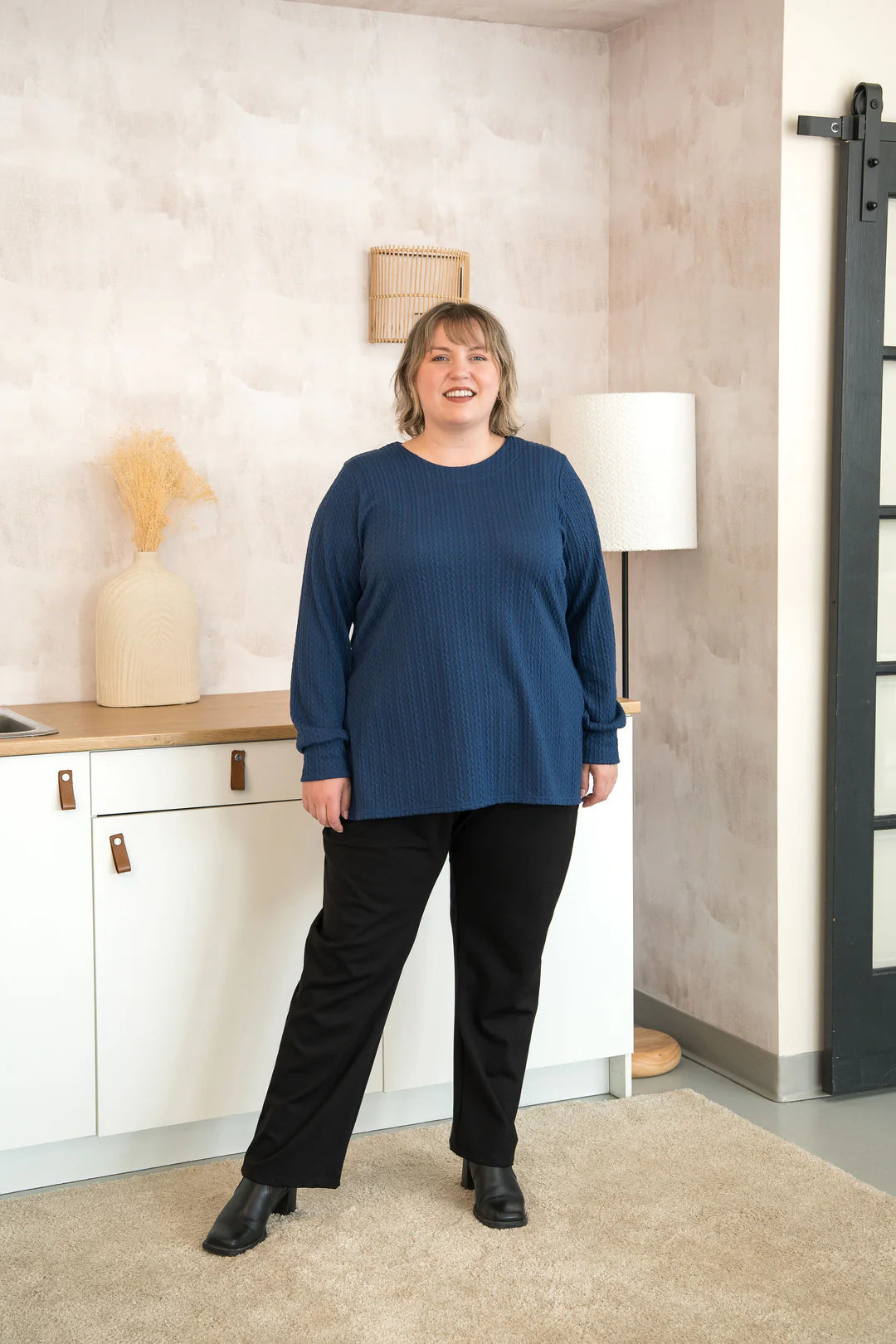 A woman wearing the Manhattan Pants by Cherry Bobin in Black straight leg Ponte di Roma pants with a pull on waist. She is wearing them with the Cosmopolitain Sweater  and standing in a room with shelves. 