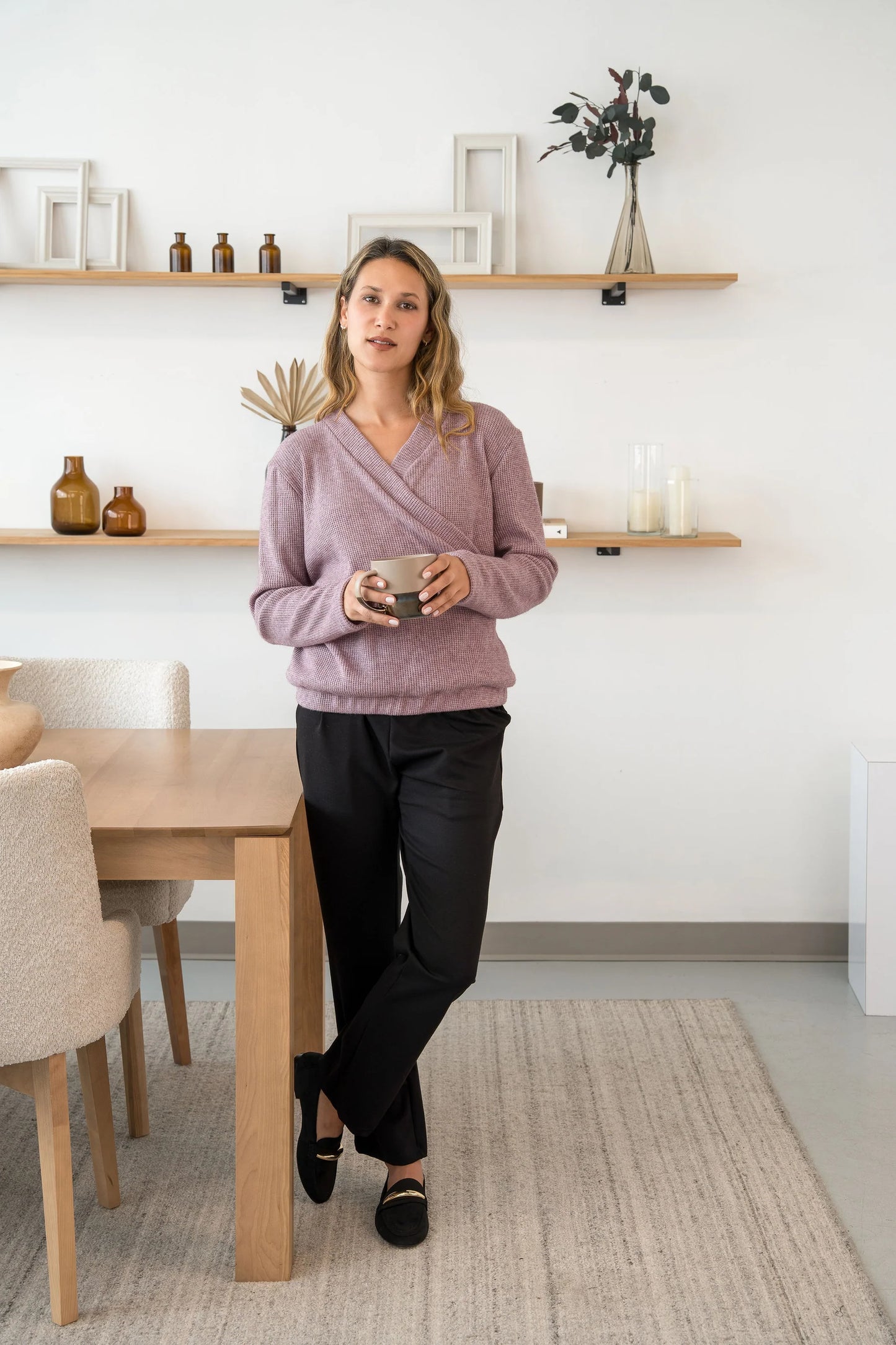 A woman wearing the Manhattan Pants by Cherry Bobin in Black, straight leg Ponte di Roma pants with a pull on waist. She is wearing them with the a mauve sweater and standing in a room with shelves. 