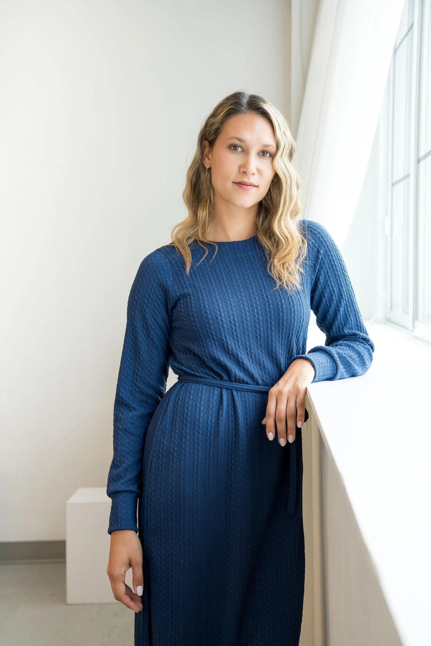 A woman wearing the Negroni Dress by Cherry Bobin in Blue, a mid-calf dress with in cable-knit textured fabric, side slits, and long, slighty puffed sleeves. She is standing near a window in a white room. 