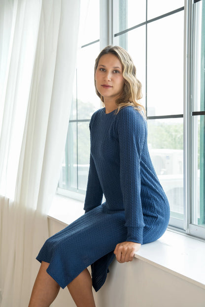 A woman wearing the Negroni Dress by Cherry Bobin in Blue, a mid-calf dress with in cable-knit textured fabric, side slits, and long, slighty puffed sleeves. She is seated in a window sill in a white room. 