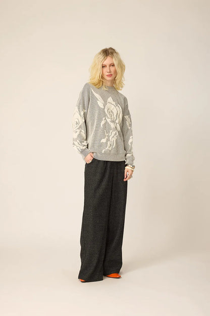 A woman wearing the Howard Sweater from Chlo by Cokluch in Natural, with a striped fabric and floral applique in an oversized cut. She is wearing it with grey pants and standing againts a beige background