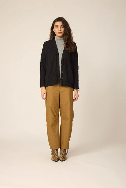 Woman wearing the Gus Cardigan from Cokluch in Black with ribbed fabric and floral embellishments on the front and sleeves. It has a relaxed it and five buttons. She is wearing it over an a striped top and ochre pants in front of a white background.