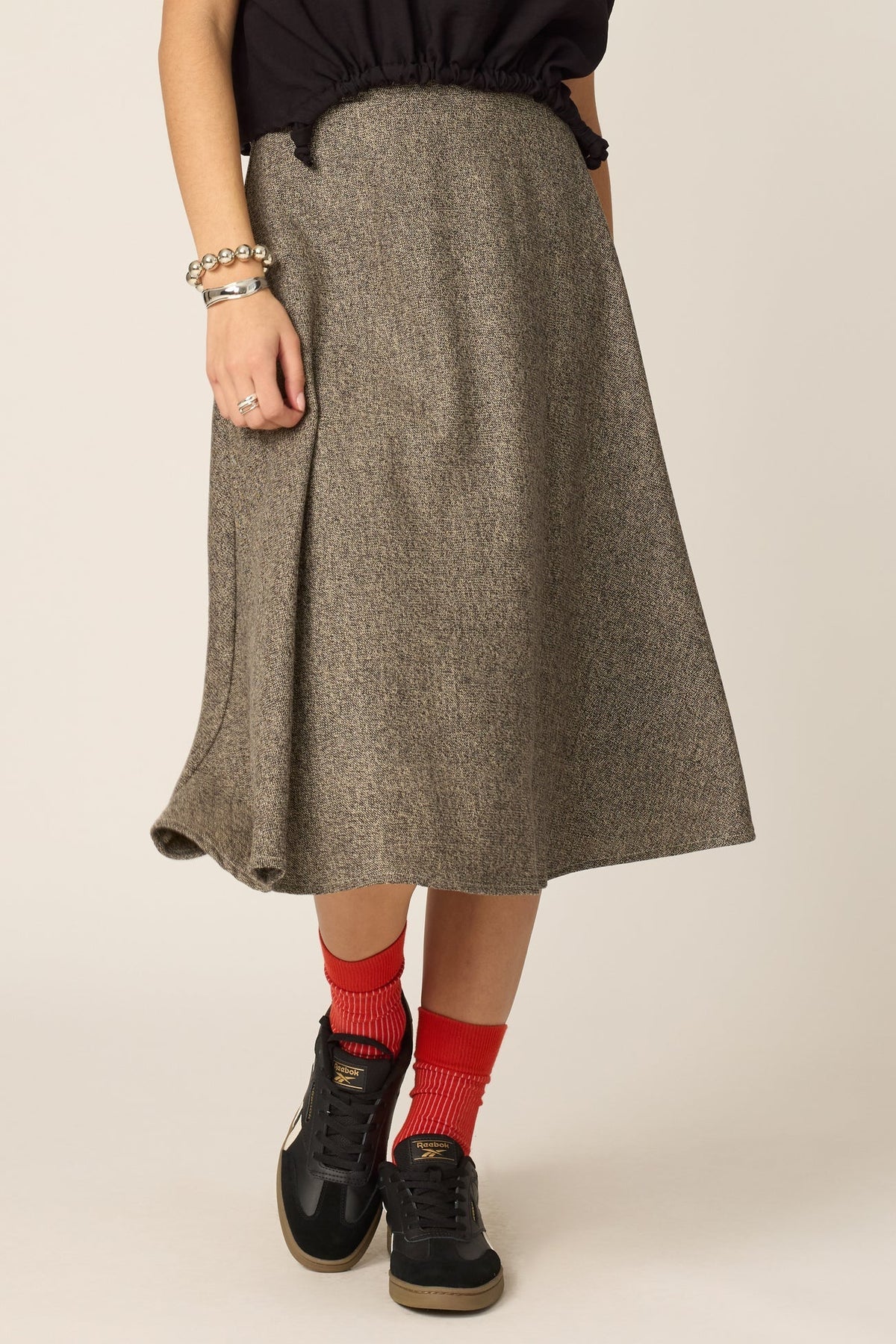 Waist down view of a woman wearing the Amber Skirt from Chlo by Cokluch in Greige Tweed, a full, flared mid-calf skirt with a high waist and adjustable side drawstrings with metal rings.  She is wearing it with a black top and standing against a grey background.