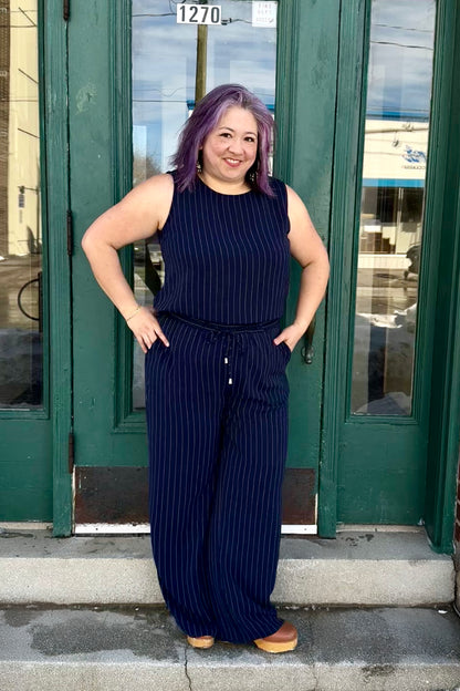A woman wearing the Ginnie Camisole by Pure Essence, a sleeveless navy/white pinstripe top with a relaxed fit. She is wearing it with the matching 
Gabby Pants and standing in front of a green door. 