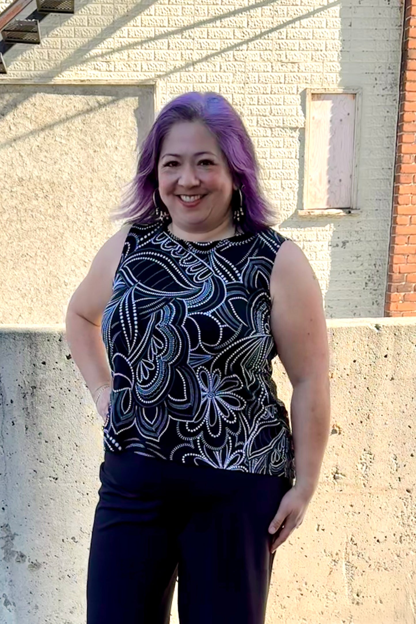 A woman wearing the Sunrise Camisole by Rien ne se Perd in Black/White floral, with a wide neck, wide straps, and a rounded hi-low hemline. She is wearing it with black pants and standing outside in front of a brick building. 
