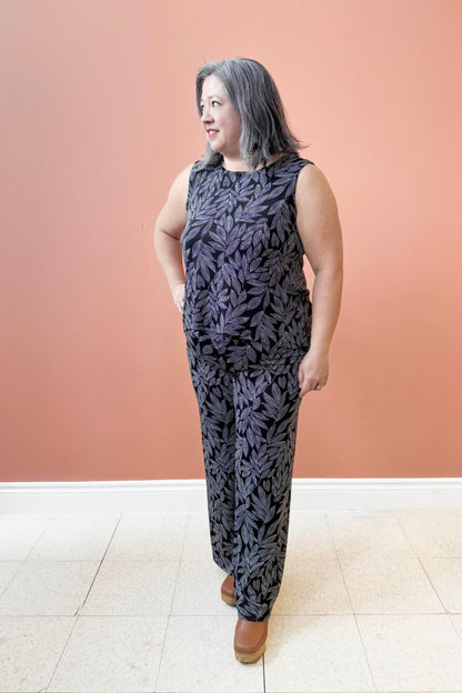 Christina is wearing the Swing Cami by Rien ne se Perd in Black and White Foliage, a trapeze cut tank with a hi-low rounded hemline. She is wearing it with the matching Rumba Palazzo pants and standing  in front of a pink wall. 