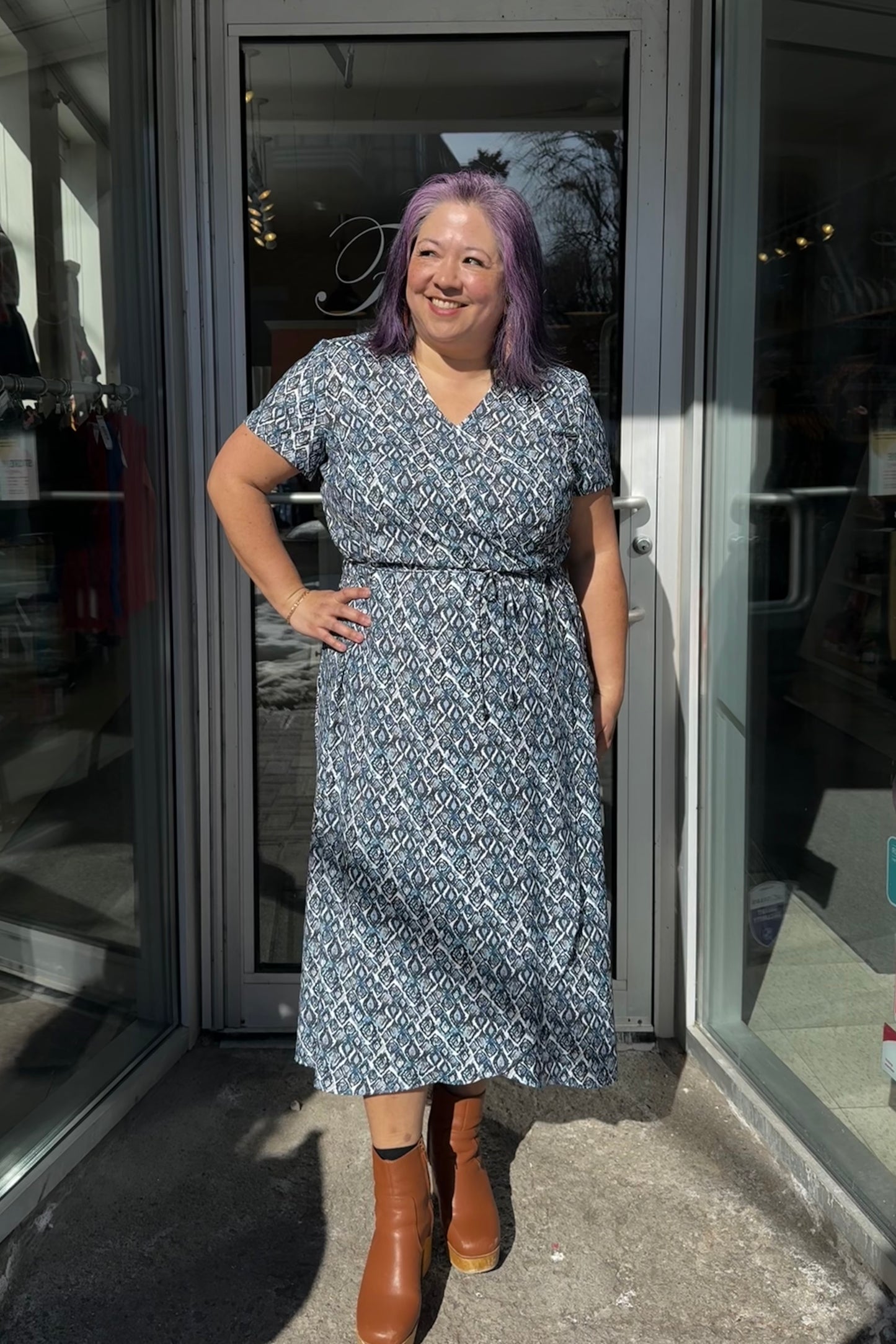 A woman wearing the Galea Dress by Pure Essence in Blue Lattice print. This faux-wrap dress has short sleeves, a gathered waist with a tie and a midi-length tulip hem. She is standing outside the Flock entrance.