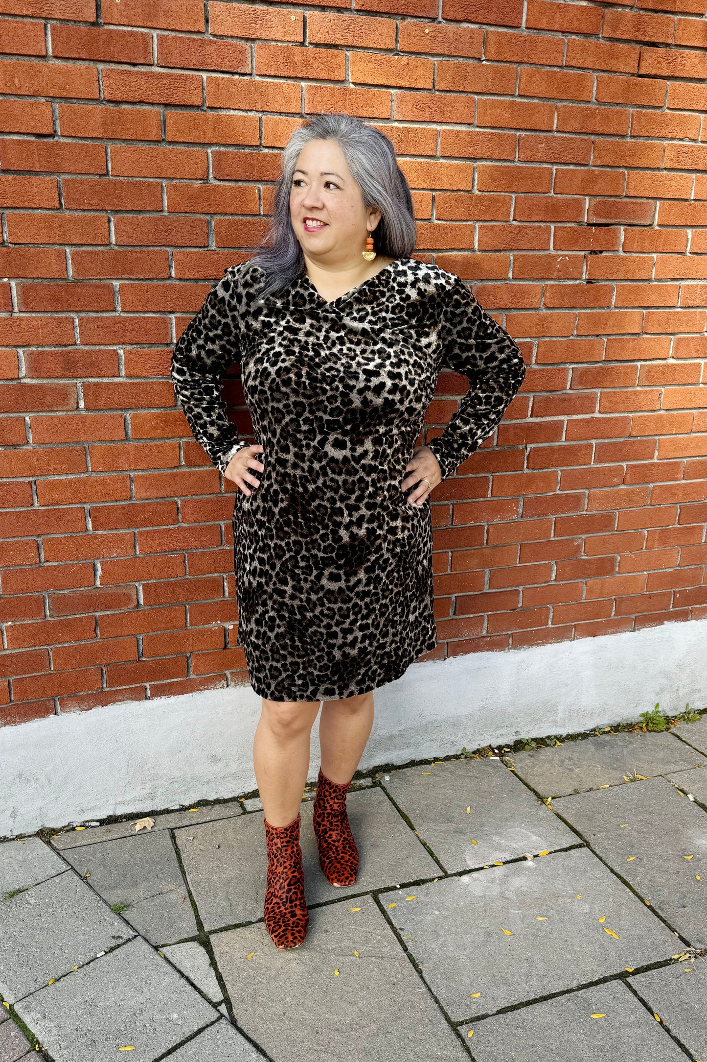 A woman wearing the Luc Fontaine Panthera Dress in Leopard Print, featuring a faux-wrap neckline, long sleeves, fuzzy animal print fabric, a sheath shape, and an above the knee length, standing in front of a brick wall. 