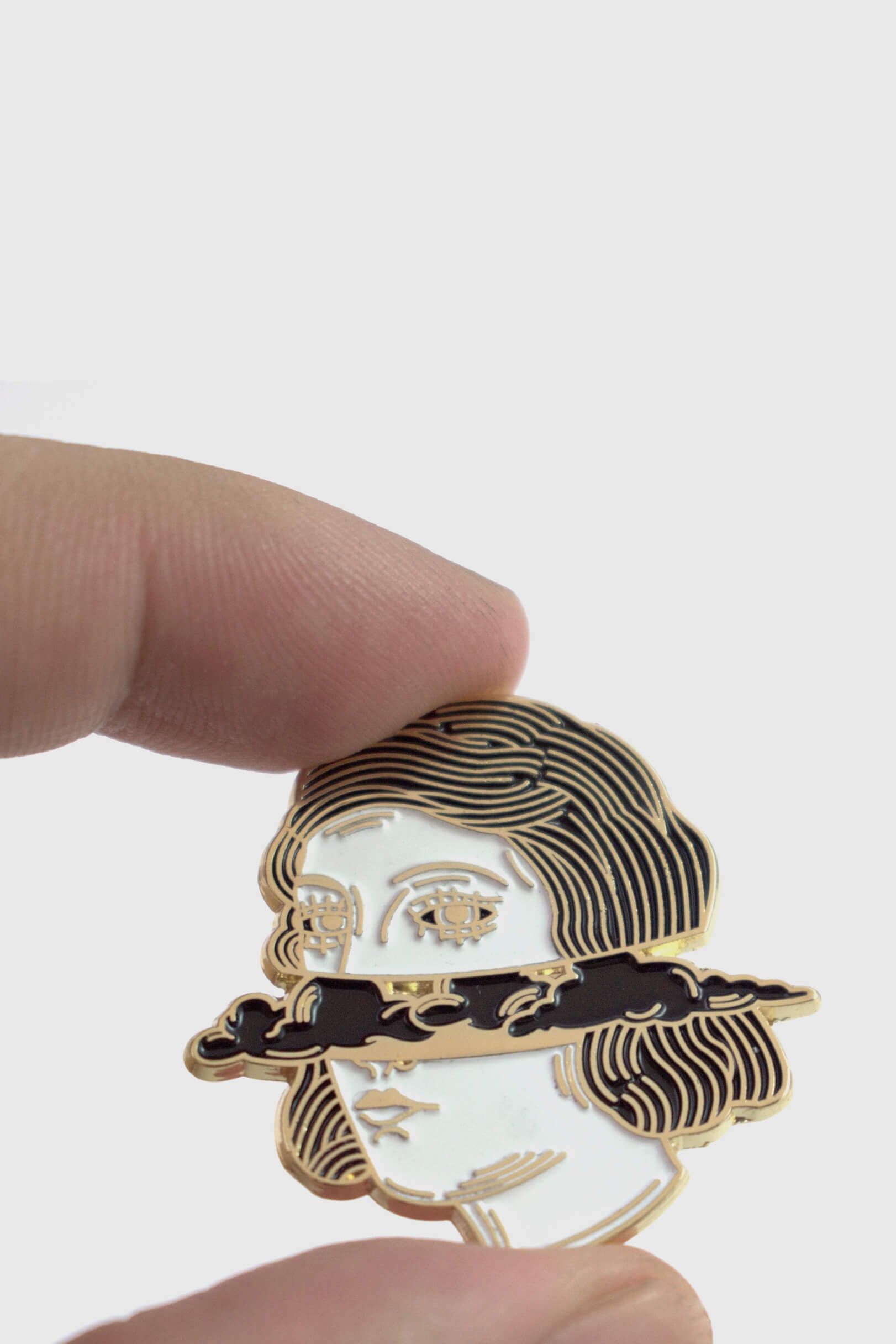 A hand holding The Cloud Head pin from Badaboöm Studio, depicting a women's face horizontally bisected by dark clouds, is shown against a white background. 
