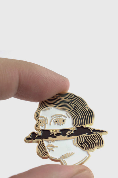 A hand holding The Cloud Head pin from Badaboöm Studio, depicting a women's face horizontally bisected by dark clouds, is shown against a white background. 