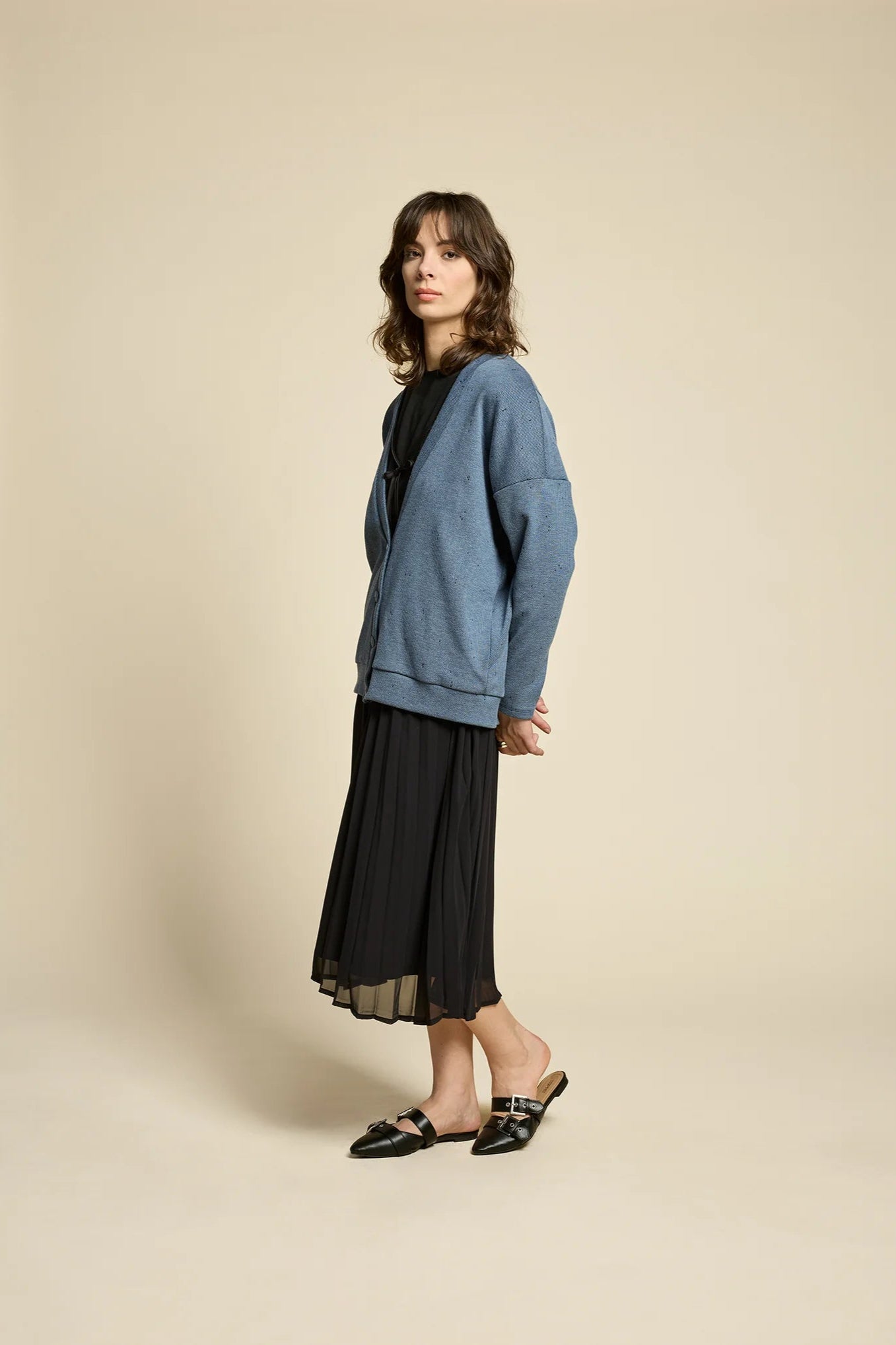 A woman wearing the Flowers Skirt by Cokluch in Black, a midi-length pleated skirt with a sheer overlayer and an elastic waist. She is wearing it with a blue cardigan and standing in front of a beige background. 