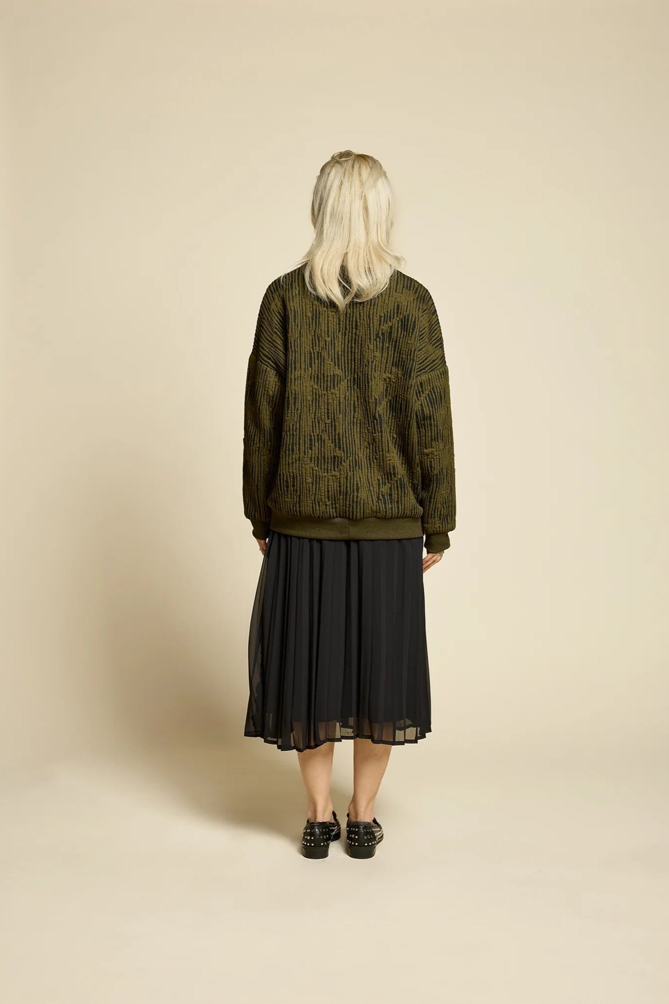 Back view of a woman wearing the Flowers Skirt by Cokluch in Black, a midi-length pleated skirt with a sheer overlayer and an elastic waist. She is wearing it with a the Hotel Sweater in Olive  and standing in front of a beige background. 