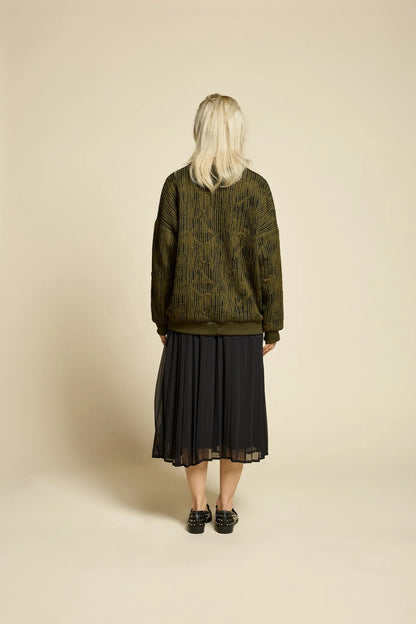 Back view of a woman wearing the Flowers Skirt by Cokluch in Black, a midi-length pleated skirt with a sheer overlayer and an elastic waist. She is wearing it with a the Hotel Sweater in Olive  and standing in front of a beige background. 