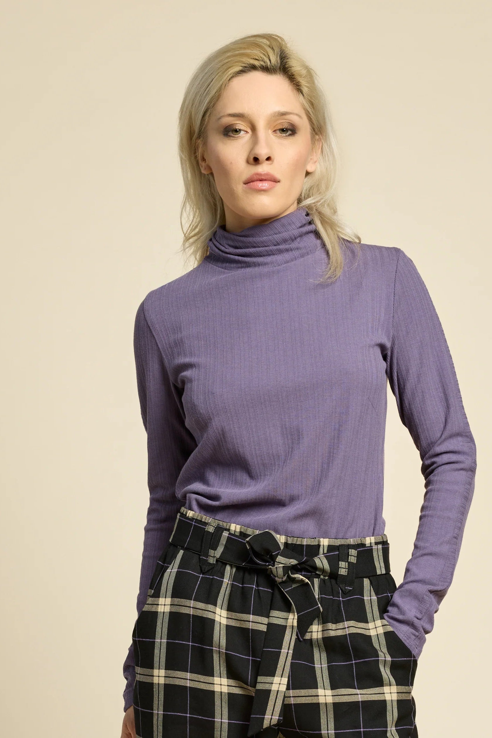 A woman wearing the Solo Top by Cokluch in Heron, a fitted turtleneck with a tone on tone striped, textured fabric and a twist detail at the neck. She is wearing it with the West Coast pants in Black Plaid and standing in front of a beige background. 