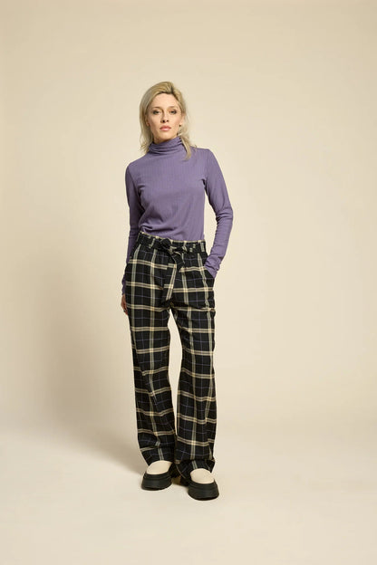 A woman wearing the Solo Top by Cokluch in Heron, a fitted turtleneck with a tone on tone striped, textured fabric and a twist detail at the neck. She is wearing it with the West Coast pants in Black Plaid and standing in front of a beige background. 