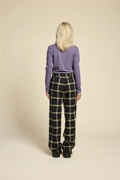 Back view of a woman wearing the Solo Top by Cokluch in Heron, a fitted turtleneck with a tone on tone striped, textured fabric and a twist detail at the neck. She is wearing it with the West Coast pants in Black  Plaid and standing in front of a beige background. 