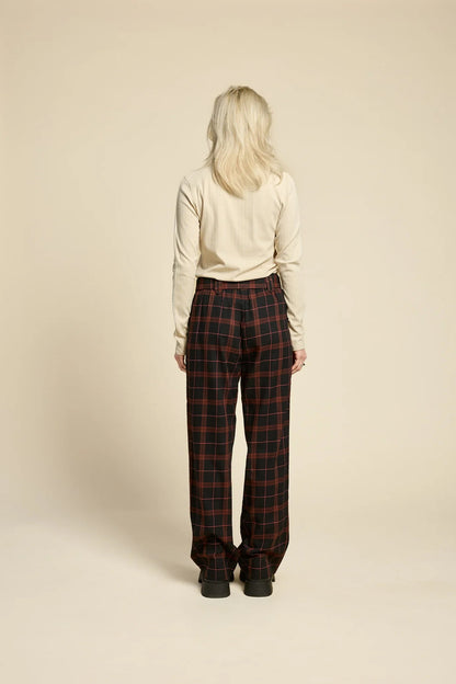 Back view of a woman wearing the Solo Top by Cokluch in Sand, a fitted turtleneck with a tone on tone striped, textured fabric and a twist detail at the neck. She is wearing it with the West Coast pants in Burgundy Plaid and standing in front of a beige background. 